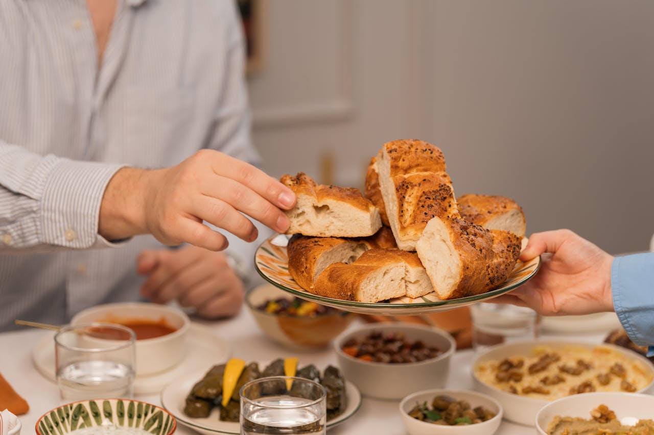 hero-img-01 Hands exchanging fresh bread at a dinner table filled with diverse dishes and drinks.