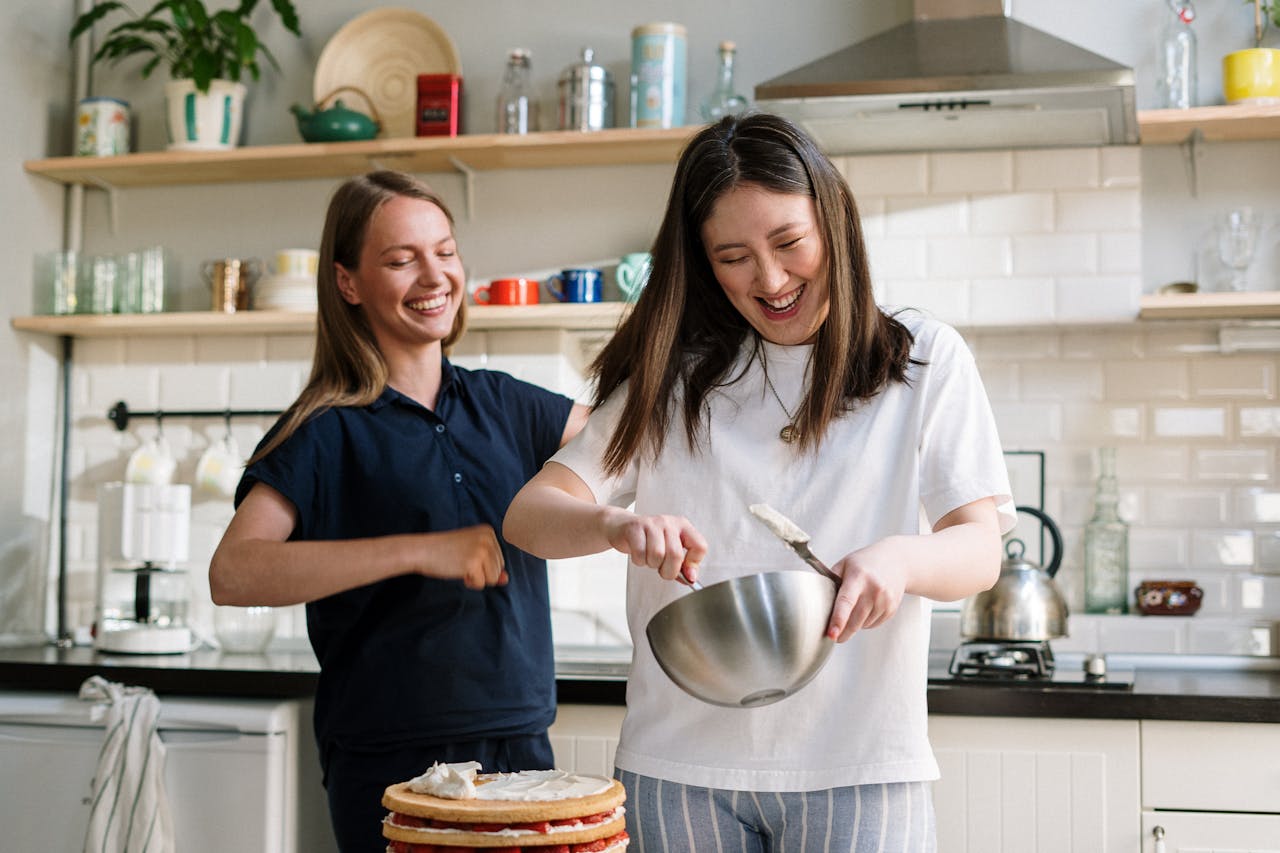 our-services-1 Two women cooking and laughing while preparing a cake in a cozy kitchen.